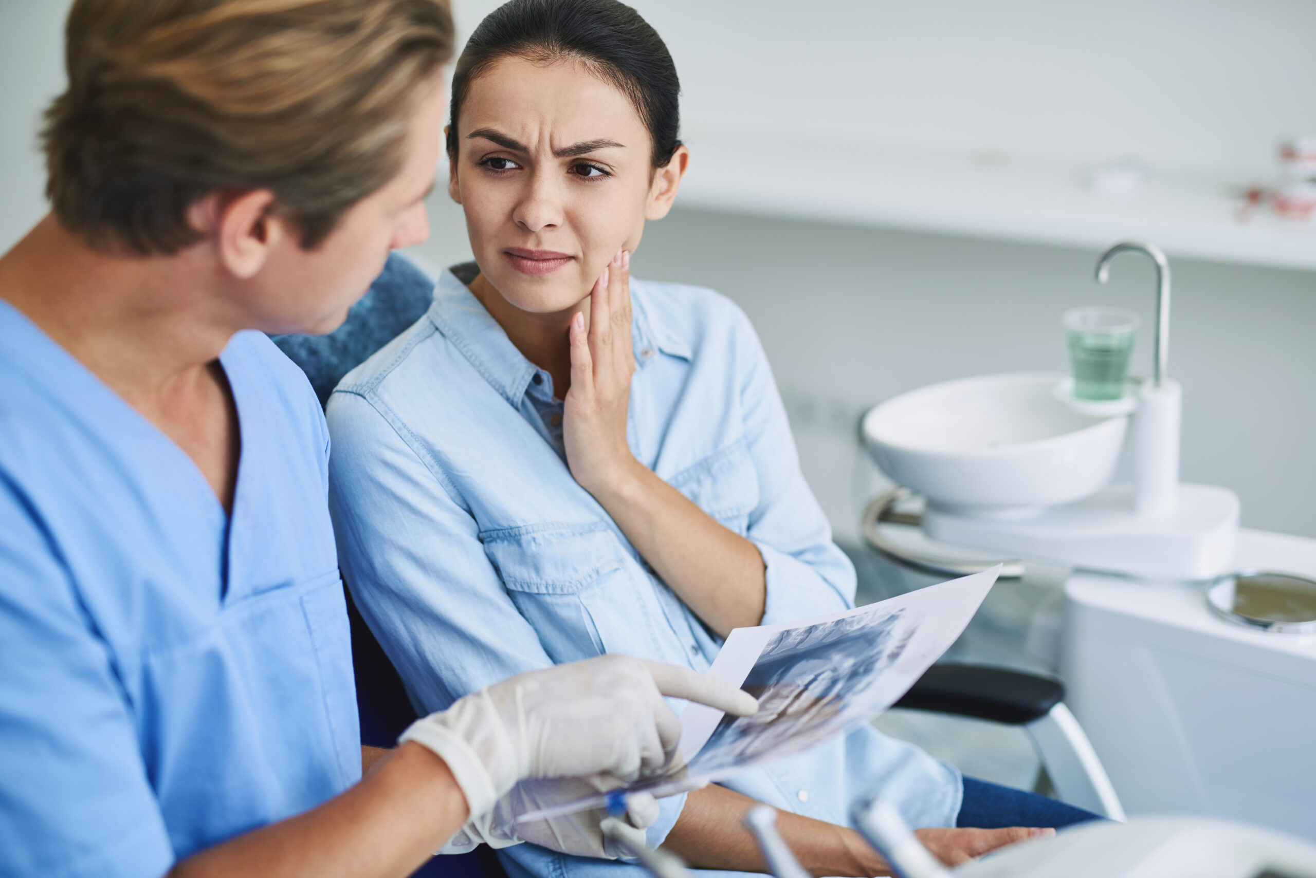 Portrait of brunette woman having toothache and touching cheek while sitting in dental chair. Stomatologist holding dental xray and pointing at it