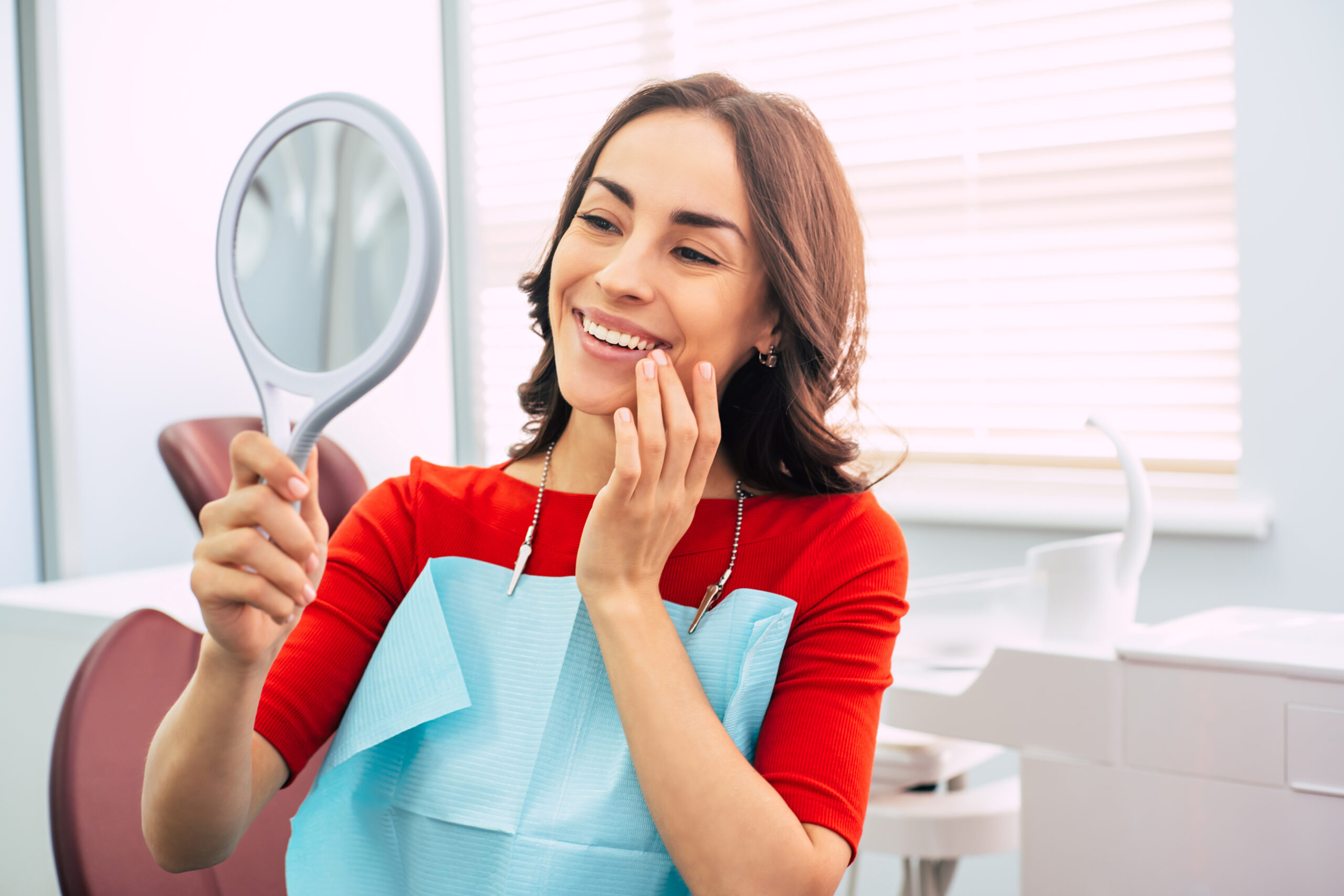 Brilliant work. Pretty girl in dentist cabinet is using a mirror to look through the ideal work of a doctor. She has a special dentist napkin over her vibrant red sweater.