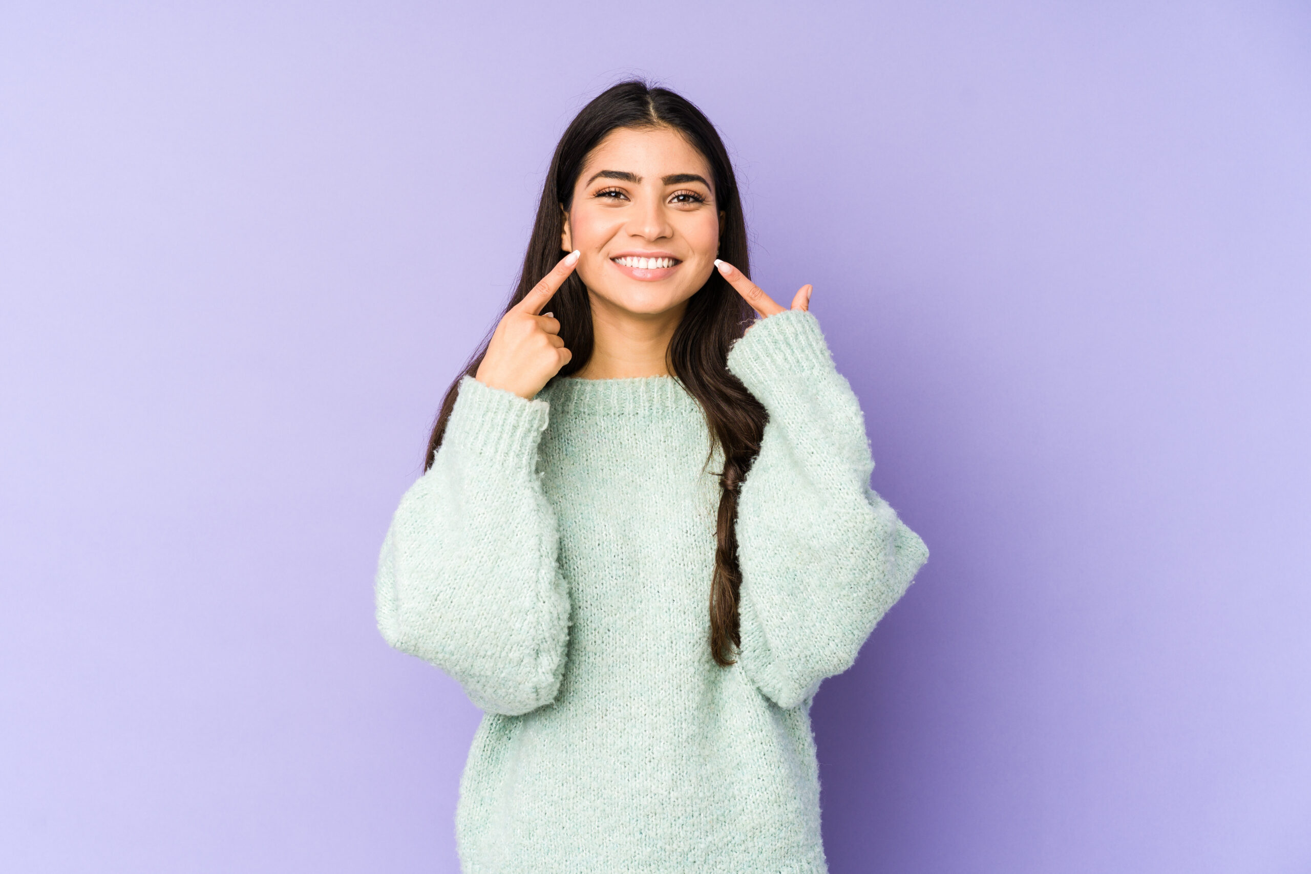 Young indian woman isolated on purple background smiles, pointing fingers at mouth.