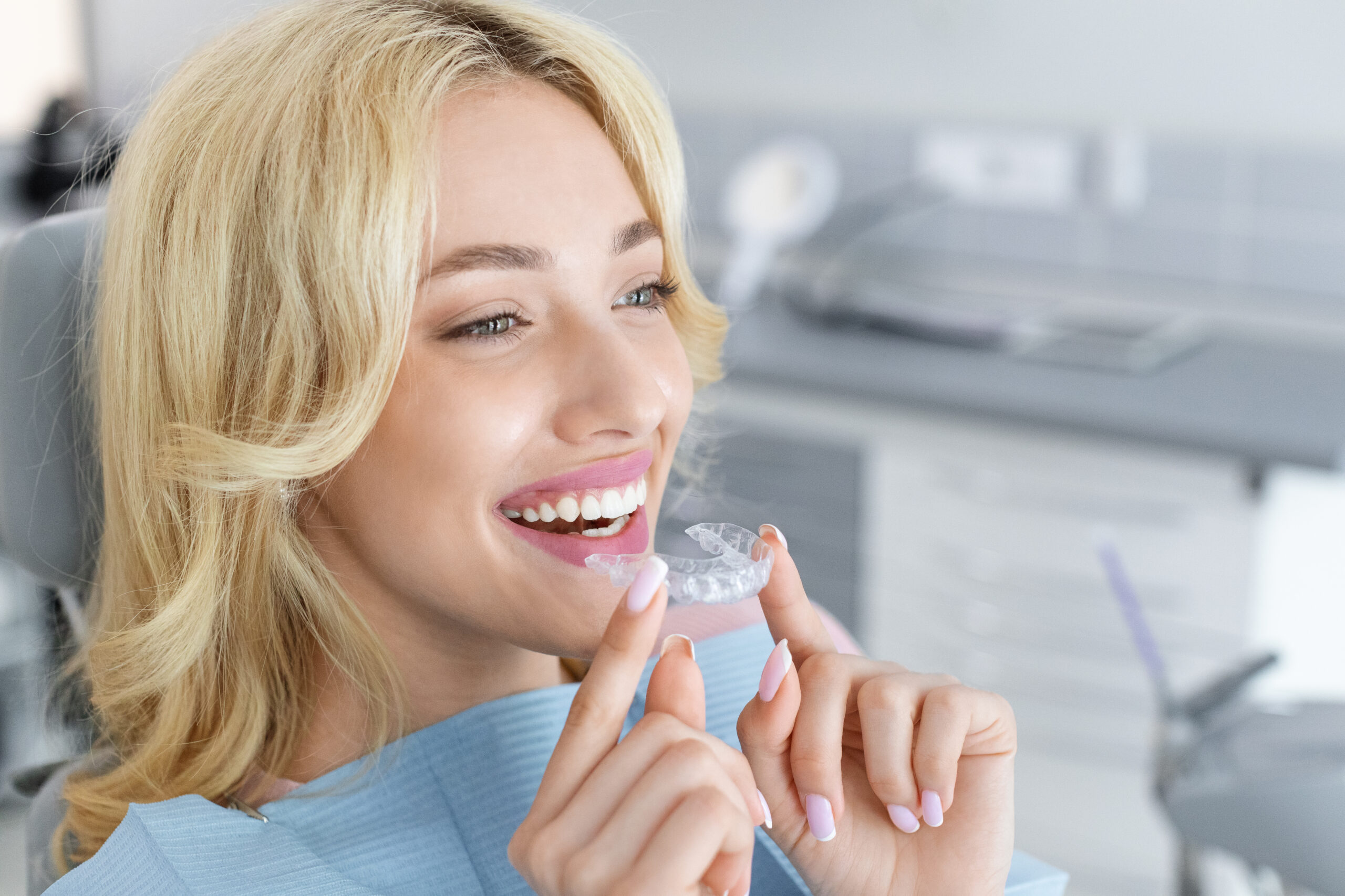 Invisalign orthodontics, modern dentistry concept. Happy young attractive blonde woman holding invisible braces or trainer while sitting at dental chair, closeup portrait, copy space