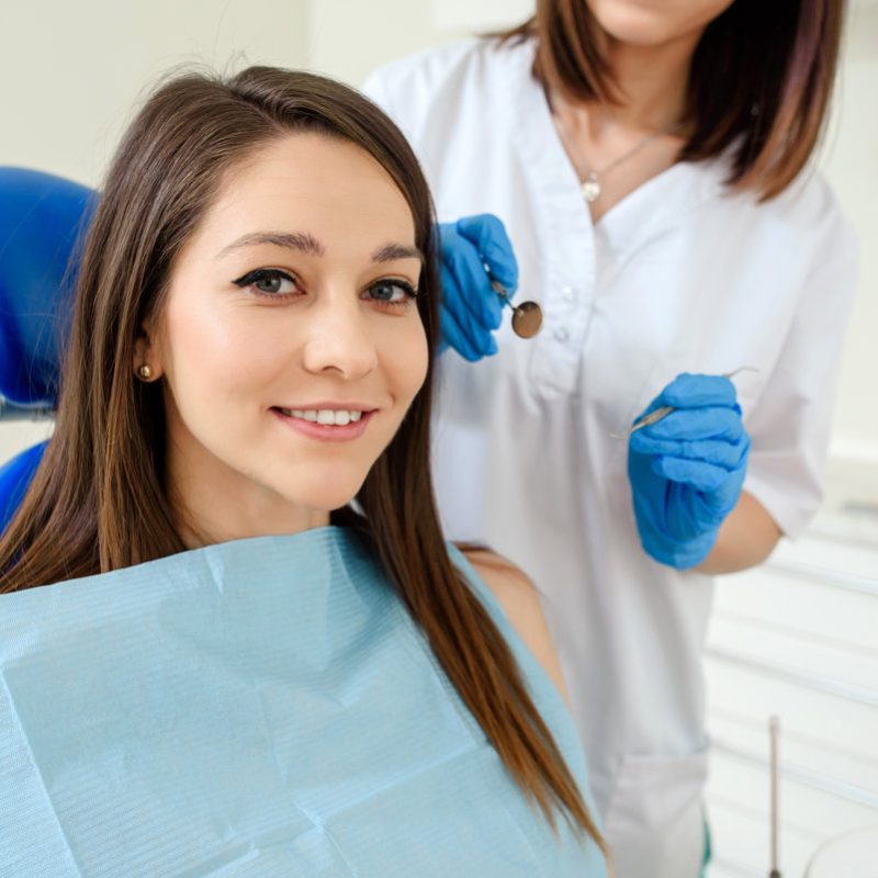 Young happy smiling woman patient in a dental clinic is receiving a dental treatment and looking at the camera. The dentist with dental equipment are behind